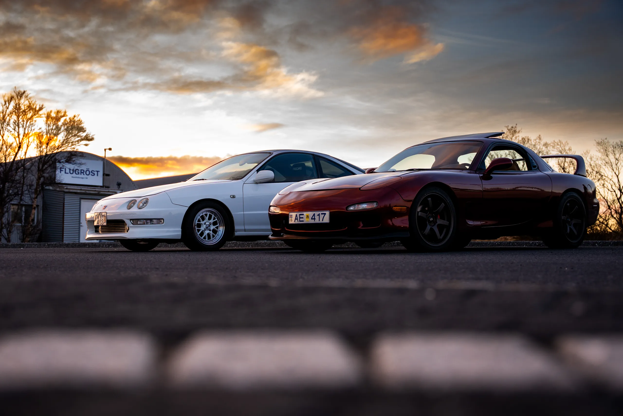 A white sports car and a burgundy sports car parked beneath a golden sunset sky