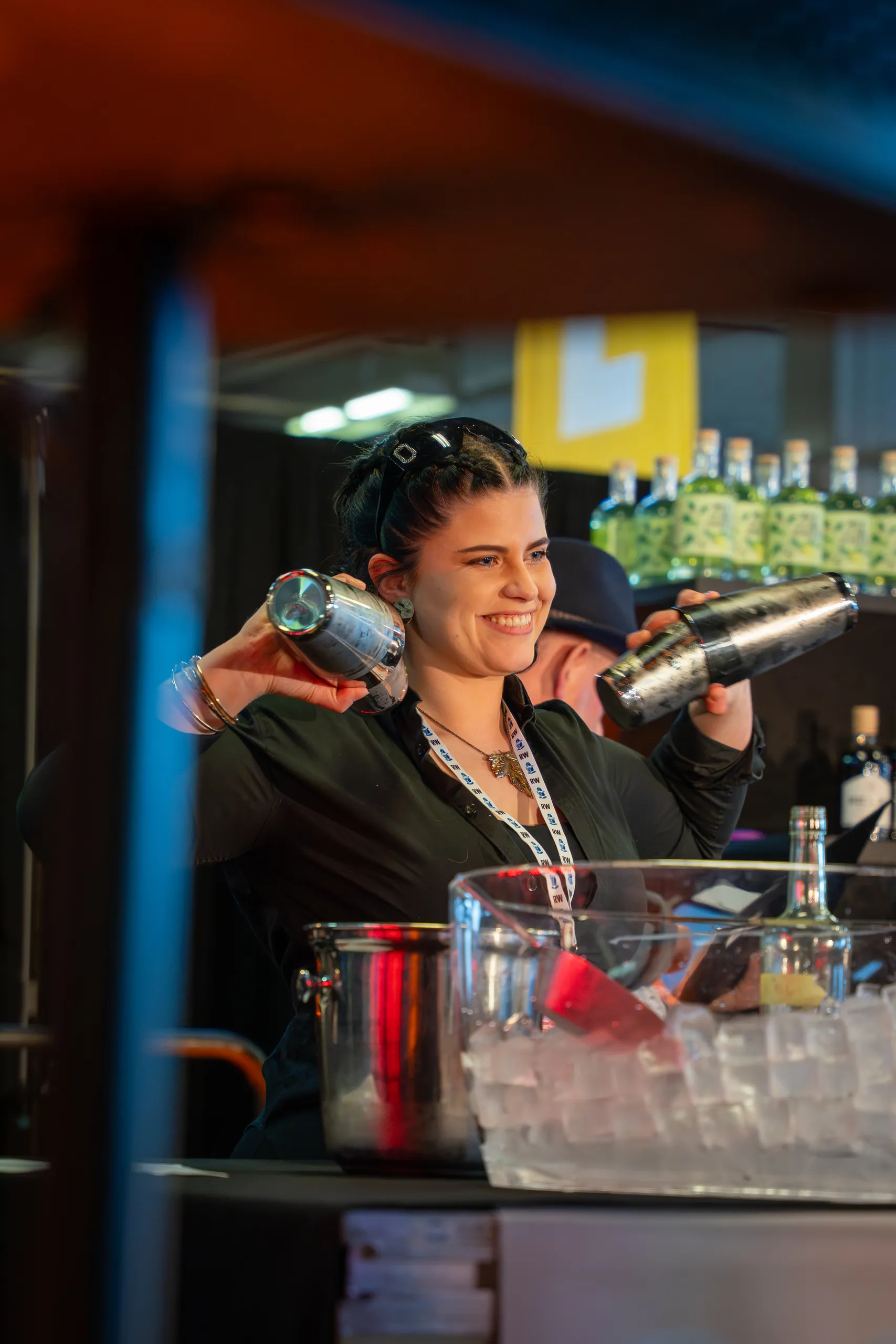 A smiling bartender holding two metal cocktail shakers behind a colorful bar