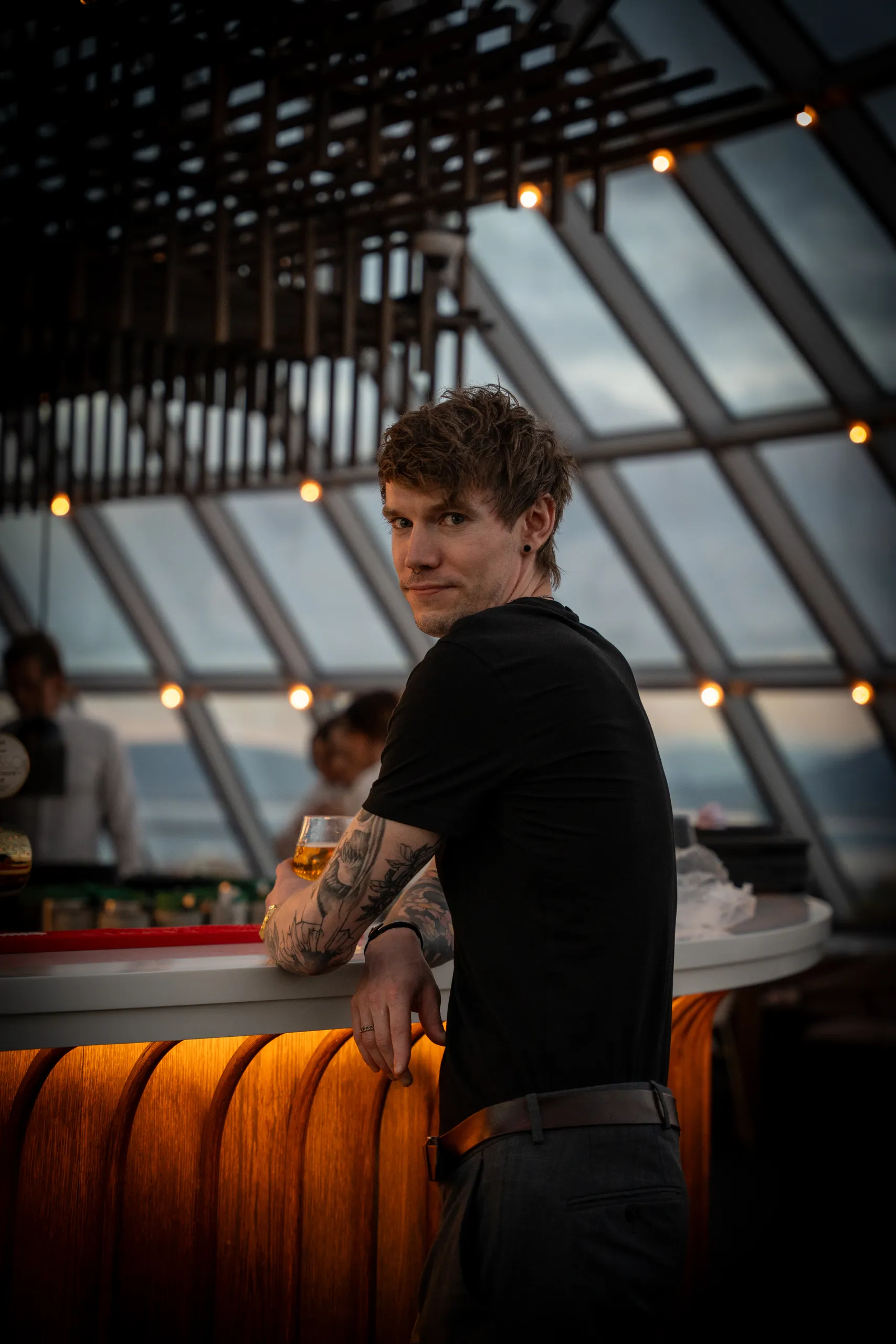 A man in a black T-shirt looking toward the camera beside a warmly lit bar