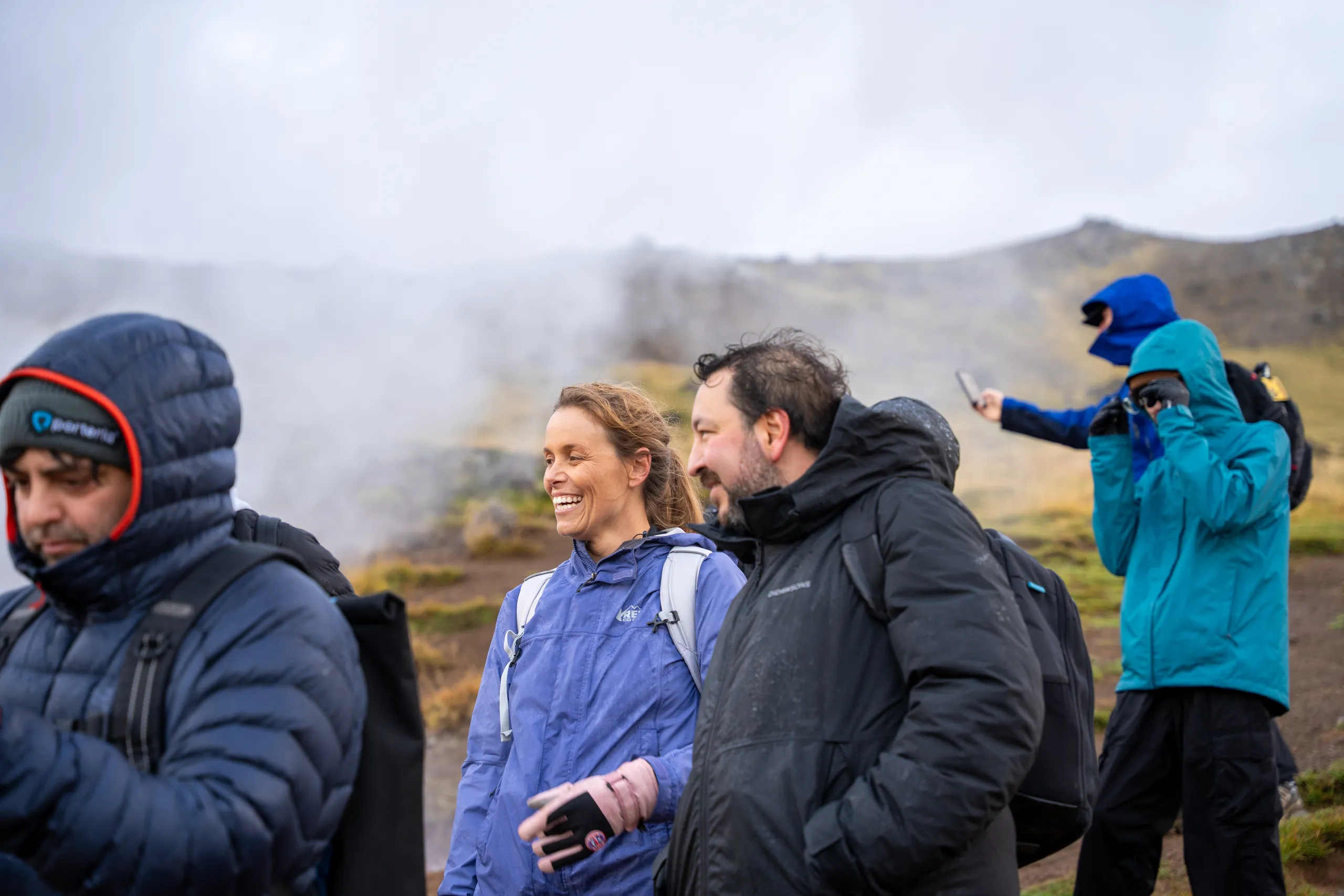 People in outdoor jackets smiling and talking beside steam rising from a rocky landscape