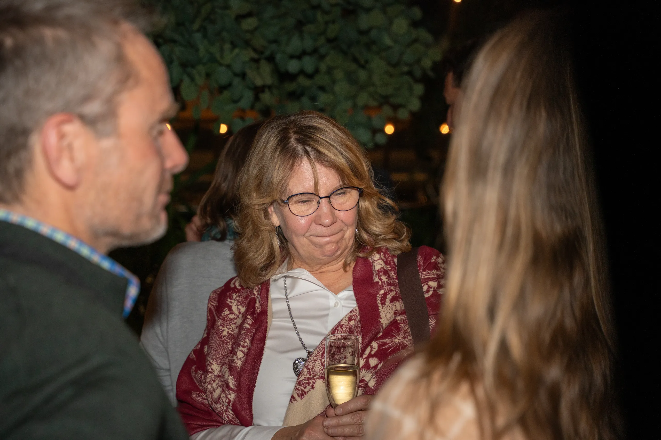 A woman with glasses smiling during a conversation at a warmly lit gathering