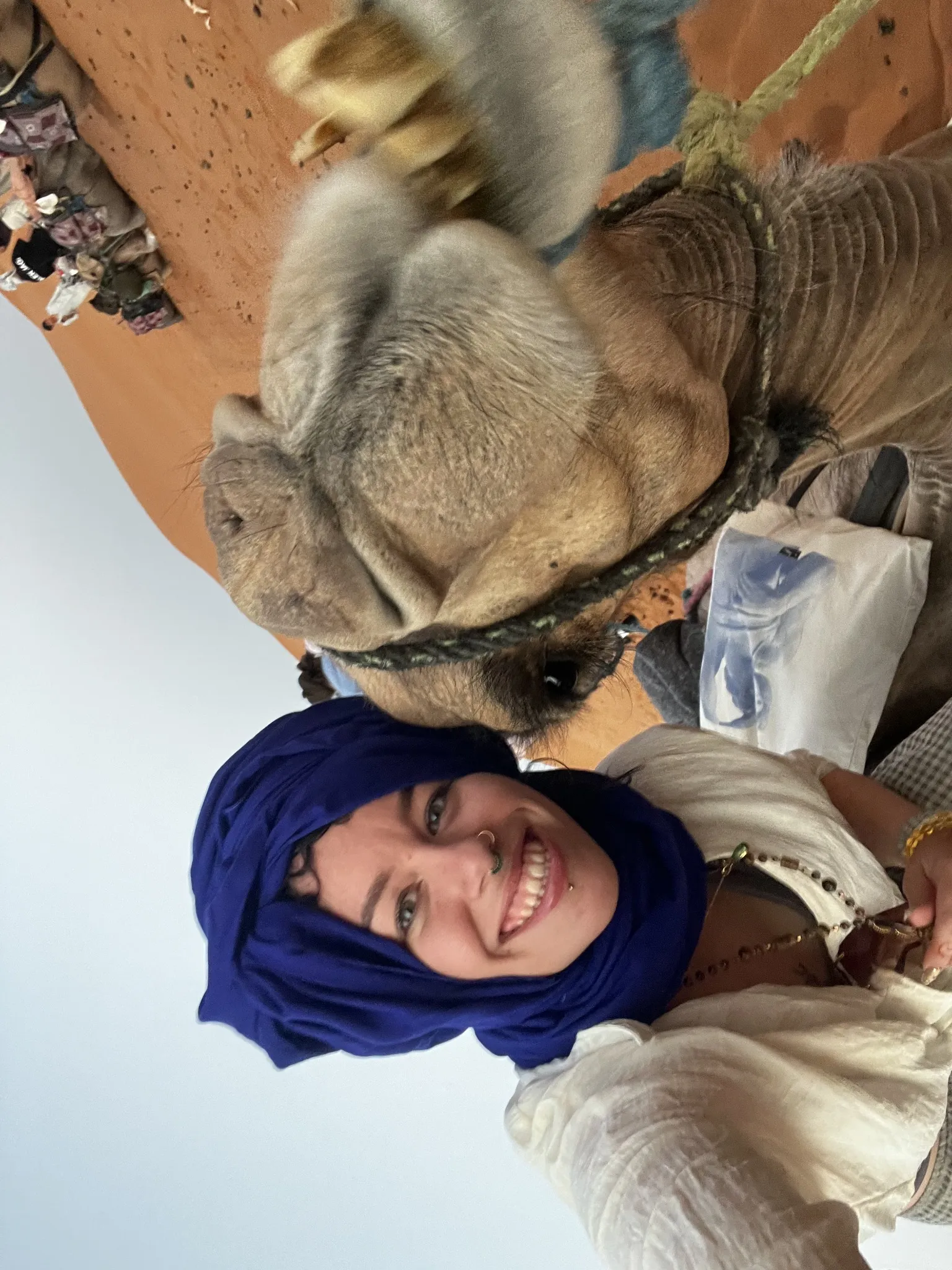 A sideways selfie of a smiling traveller in a blue headscarf beside a camel